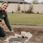 A woman kneels in muddy soil next to a large puddle, pointing at an open trench with gravel and a pipe for clay soil