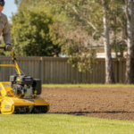 Landscaper pushing a yellow rotary hoe across bare earth during a lawn renovation in Kambah with residential homes behind